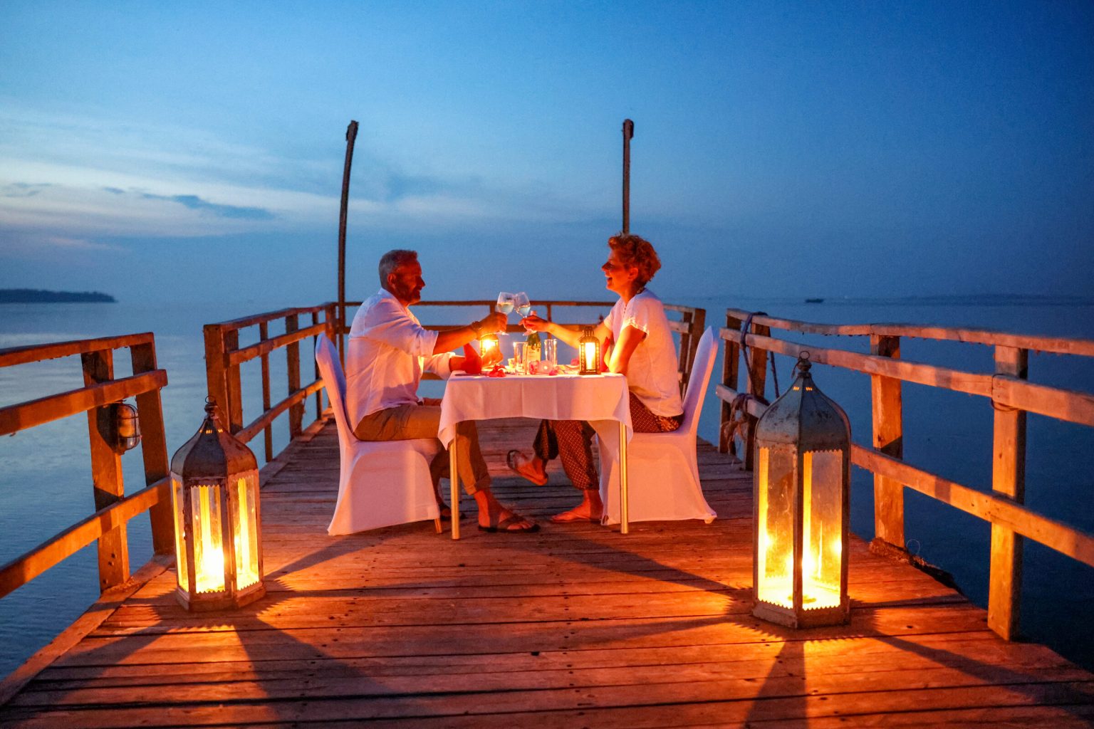 A man pours wine into a glass at a candlelit dinner table on a wooden jetty, with lanterns glowing on the deck and the lake fading into dusk behind.