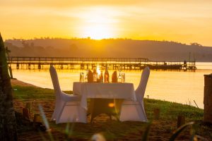 A table for two dressed in white linen stands on the sand at the water's edge as the sun sets behind a long wooden jetty on the lake.