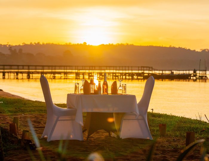 A table for two dressed in white linen stands on the sand at the water's edge as the sun sets behind a long wooden jetty on the lake.