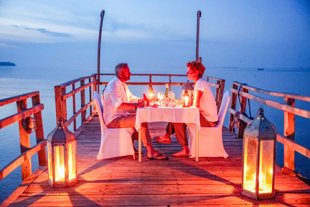 A man and a woman sit facing each other at a white-clothed table on a wooden jetty at dusk, with tall lanterns burning on the deck and the lake stretching out behind them.