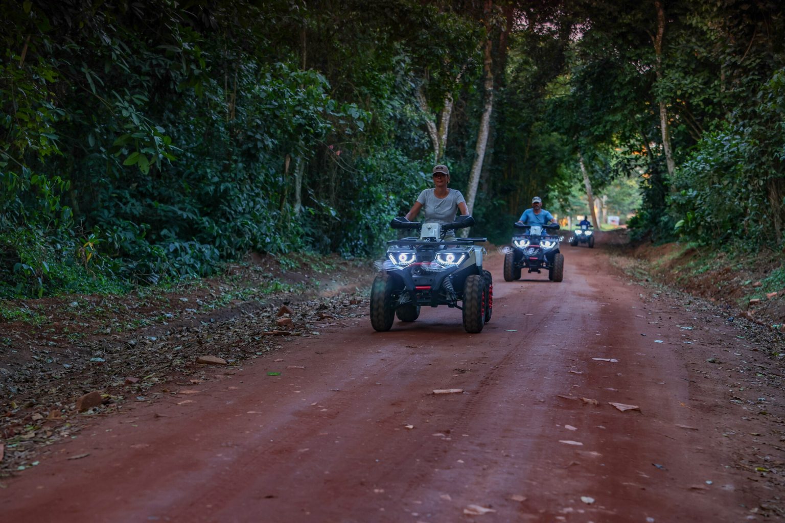 Three quad bikes with headlights on ride in single file along a red-earth track cut through dense green forest.
