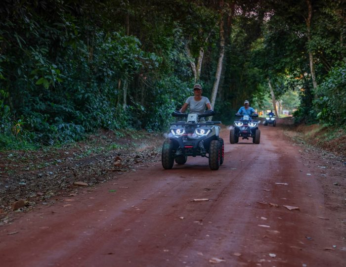 Three quad bikes with headlights on ride in single file along a red-earth track cut through dense green forest.
