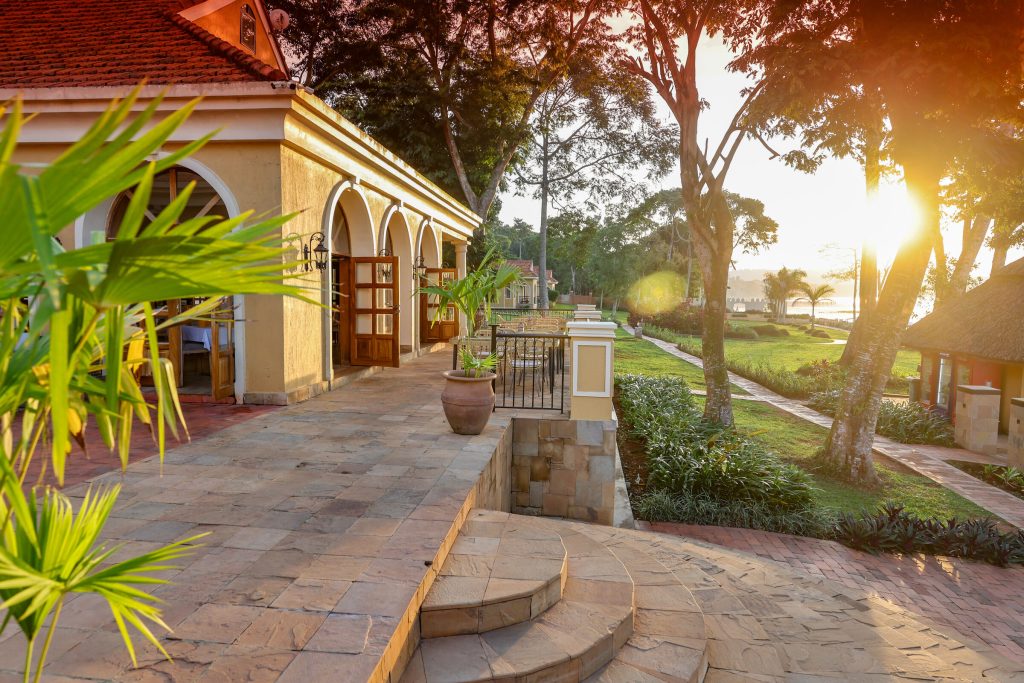 An arched colonnade beside a broad flagstone terrace catches low golden sunlight, with lawns, mature trees and the lake beyond.