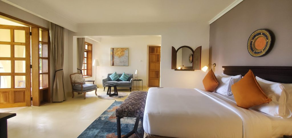 A large hotel room with a king bed dressed in white linen and orange cushions, a grey sofa and armchair seating area, and French doors opening to daylight.