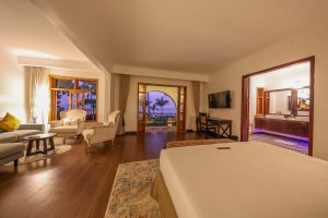 A very large room combines a seating area with sofa and armchairs, a bed in the foreground and an open bathroom to the right, with balcony doors to the lake in the centre.