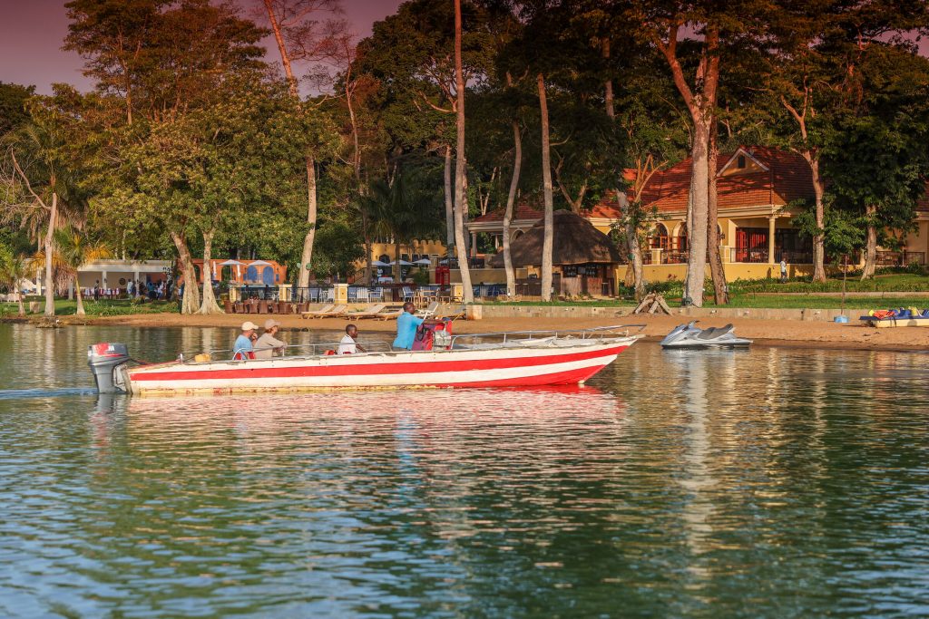 A long red and white speedboat with passengers aboard floats just off a sandy shore backed by tall trees, lodge buildings and a thatched shelter, with jet skis drawn up on the beach.