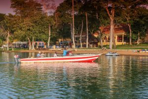 A long red and white speedboat with passengers aboard floats just off a sandy shore backed by tall trees, lodge buildings and a thatched shelter, with jet skis drawn up on the beach.