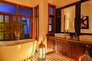 A warmly lit bathroom at night with a freestanding oval bath beside an open window, twin white basins on a dark stone counter, a Moroccan lantern on a chequered tiled floor.