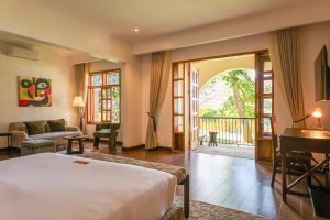 A spacious suite with a seating area, writing desk and open double doors leading onto an arched balcony among trees.
