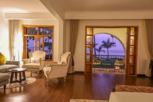 A sitting room with pale upholstered armchairs and a sofa on a polished timber floor, with tall doors opening to an arched balcony and a violet dusk sky over the lake.