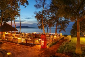 A thatched bar and a terrace of tables lit with strings of warm lights overlook the calm lake, with palms in the foreground and a pale sunset behind distant hills.