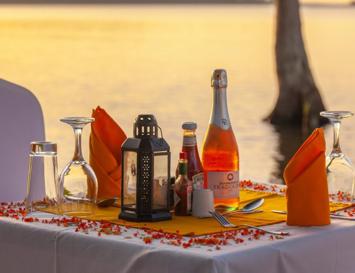 A dinner table at the water's edge laid with orange napkins, a lantern and a bottle of sparkling rose, scattered with red petals as the sun sets over the lake.