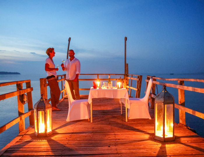 Two guests stand with drinks on a wooden jetty at blue hour, lanterns burning on the deck beside a table set for dinner.