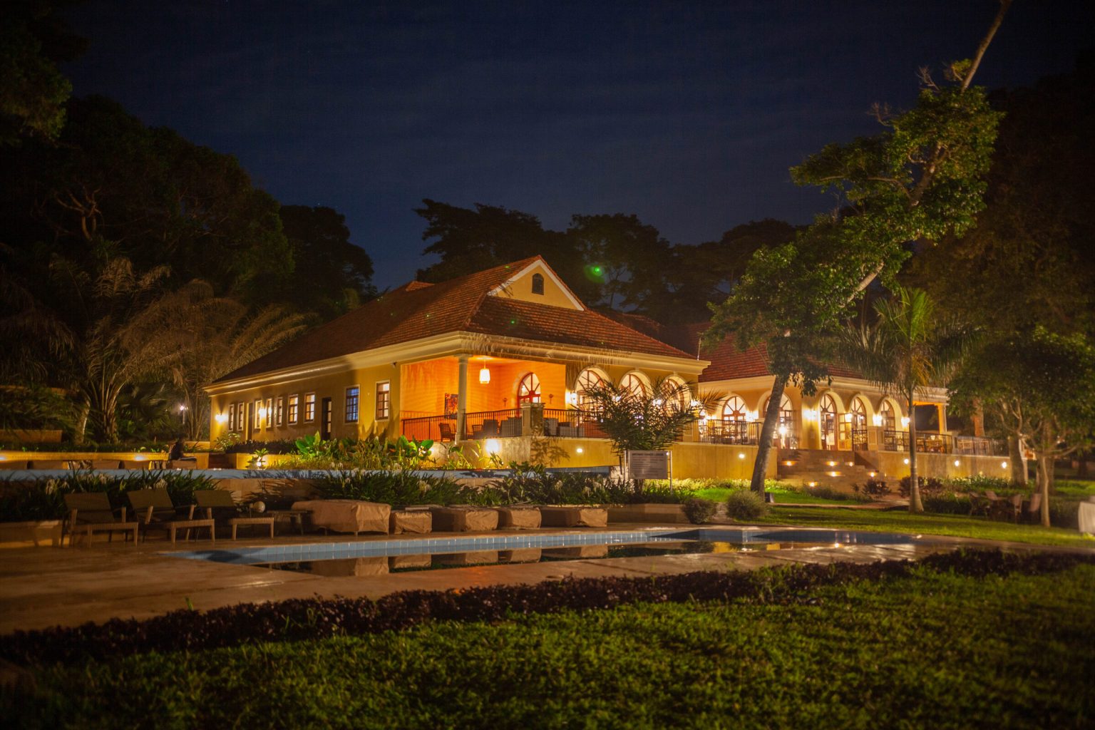 The lodge's swimming pool reflects the warm lights of the arched restaurant building above it, framed by dark palms and lawns under a night sky.