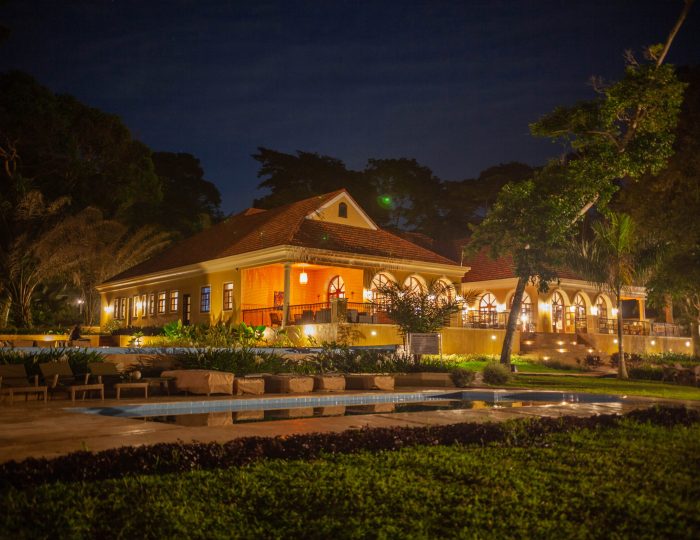 The lodge's swimming pool reflects the warm lights of the arched restaurant building above it, framed by dark palms and lawns under a night sky.