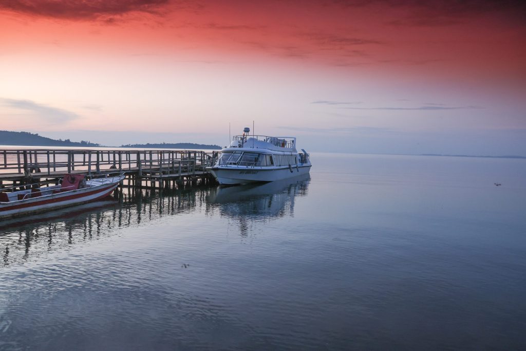 A long wooden jetty stretches into a calm lake where a white motor boat and a red fishing canoe are moored under a deep red sky.