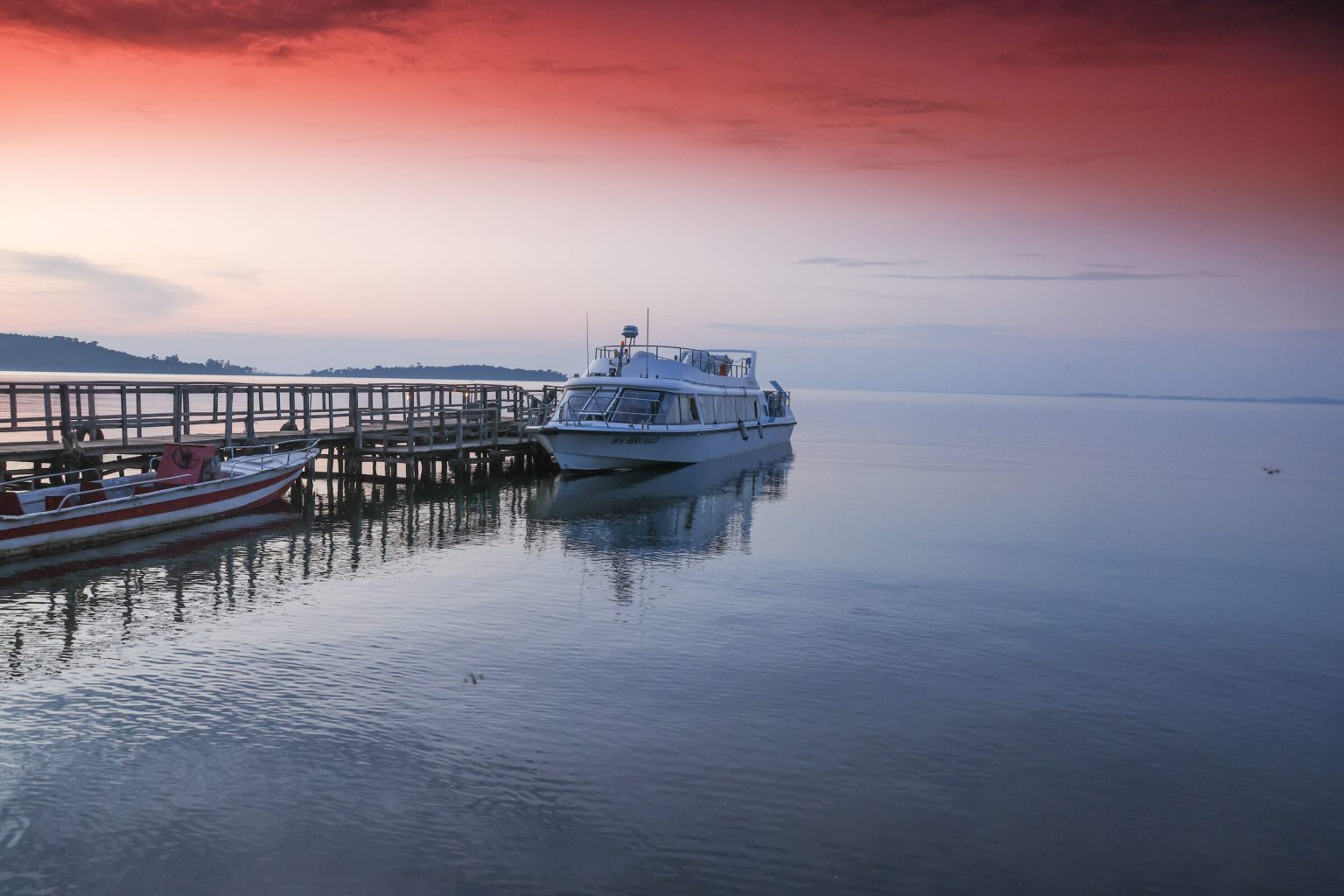 A long wooden jetty stretches into a calm lake where a white motor boat and a red fishing canoe are moored under a deep red sky.