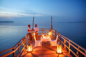 A wooden jetty runs out into a vast calm lake at dusk, with two small figures standing at the far end beside a lantern-lit dinner table.