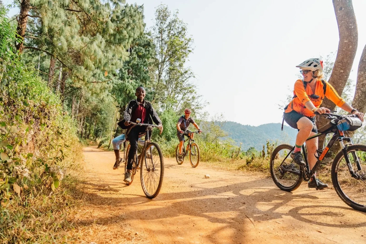 Three cyclists ride along a red dirt road lined with trees, with wooded hills in the distance.