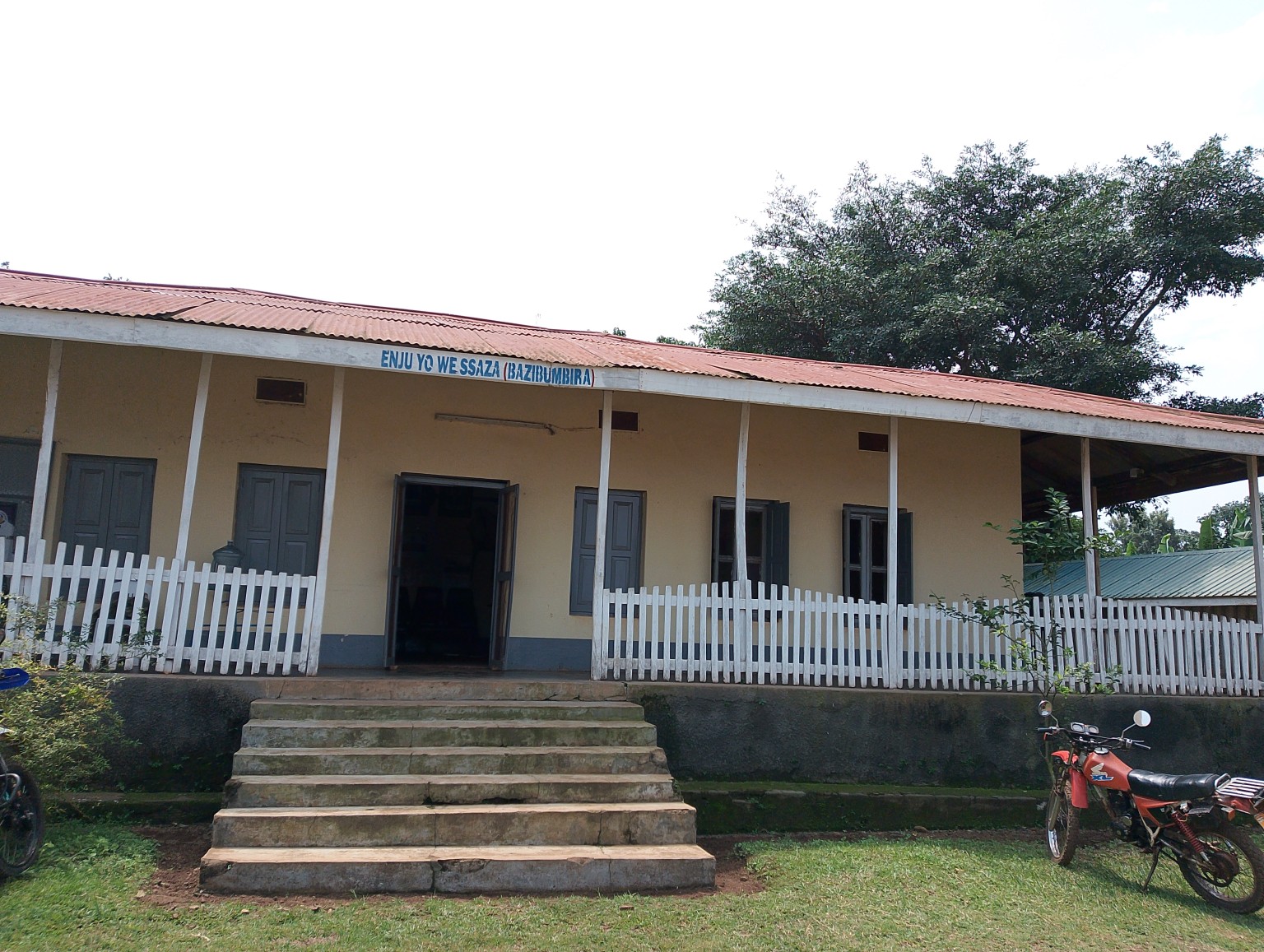 The Ssaza county headquarters at Kalangala: a long single-storey building with a red tin roof and a white picket fence, with motorcycles parked on the grass outside.